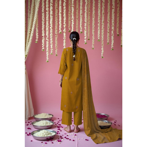 Person in a yellow traditional outfit standing in front of a pink wall with decorative elements.